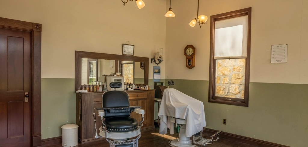An empty traditional barbershop room with a single chair, a mirrored counter, a wall clock and a tall window